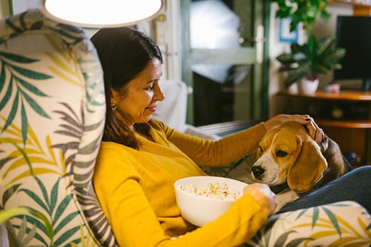 Woman in yellow sweater sits in a chair with a bowl of popcorn and a beagle in her lap.