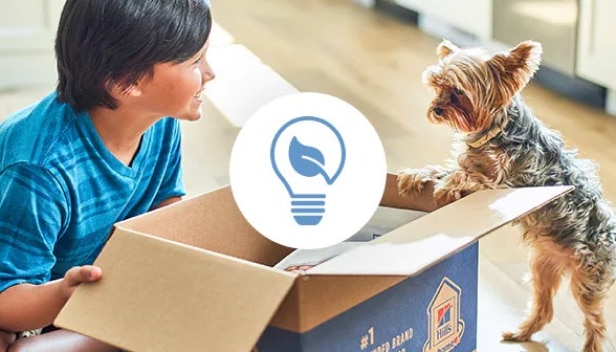 Un jeune garçon et un chien regardant dans une boîte Hill’s recyclable  sur laquelle figure une icône de réduction des déchets